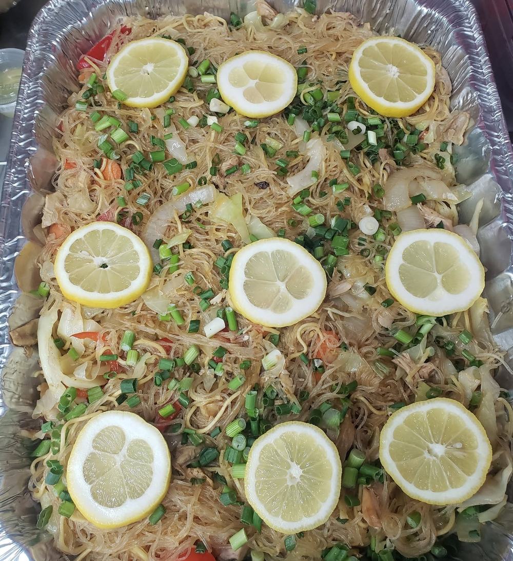 A big foil party tray of pancit noodles topped with green onions and lemon slices